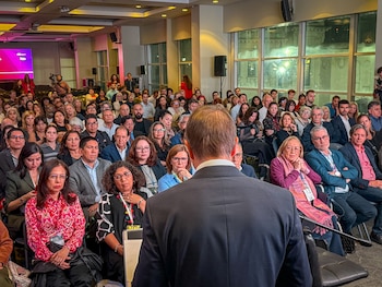 Vista traseira do palestrante em frente a uma sala cheia de pessoas sentadas, observando e ouvindo atentamente. Há uma grande janela nos fundos.