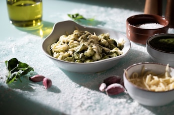 Un cuenco blanco con ñoquis, pesto y queso rallado rodeado de recipientes con sal, salsa y queso, además de ajo, albahaca y aceite