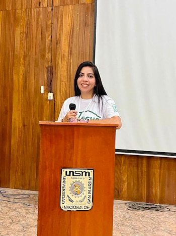 Mujer joven sonriendo con micrófono en un podio con el escudo de la Universidad Nacional de San Martín y Tarapoto, con pared de madera y pantalla blanca de fondo