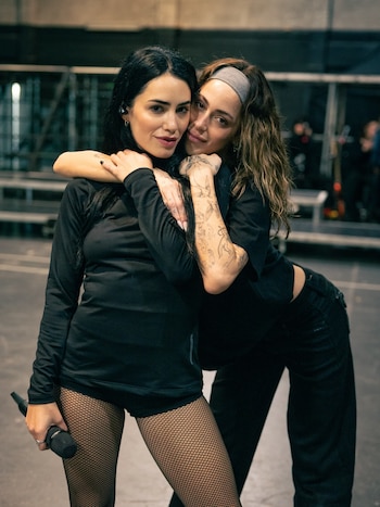 Lali Espósito viste de negro y sostiene un micrófono mientras otra mujer con tatuajes en el brazo la abraza por el cuello en un escenario