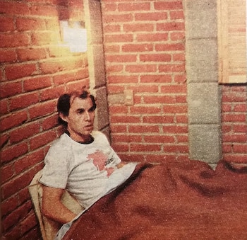 Pintura de un hombre sentado o reclinado contra una pared de ladrillo rojo, vestido con una camiseta blanca con diseño rojo y cubierto por una manta marrón rojiza