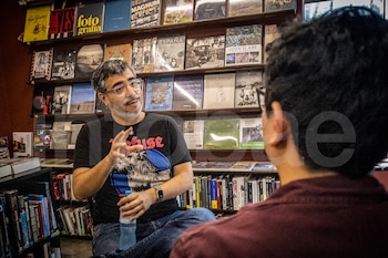 Hombre con gafas, barba y camiseta oscura gesticula, sosteniendo una botella. Espalda de otra persona. Estanterías con libros en el fondo