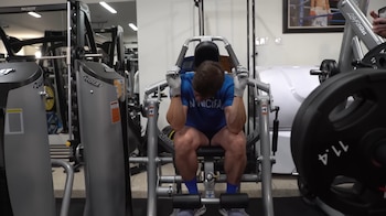 Mark Wahlberg, de frente, sentado en una máquina de abdominales en un gimnasio. Viste una camiseta azul, pantalones cortos oscuros, guantes blancos y calcetines azules