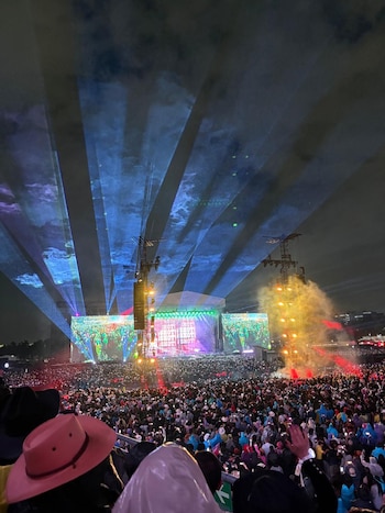 Multitud en concierto nocturno con escenario grande, pantallas luminosas, luces láser azules y columnas de humo o vapor de agua rojo-naranja