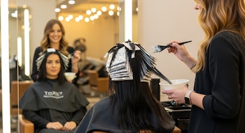 Una mujer sentada de espaldas con el cabello envuelto en papel de aluminio, mientras una estilista le aplica decolorante con una brocha en un salón.