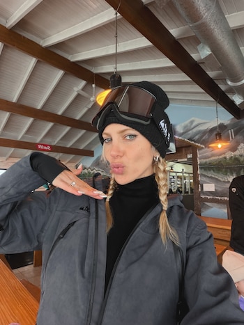 Cande Ruggeri con gorro negro, gafas de esquí y chaqueta gris posa con una mano levantada dentro de una cabaña