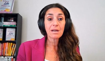 Una mujer de cabello oscuro con auriculares negros y saco rosa habla ante la cámara con un estante con libros al fondo