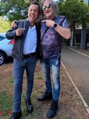 Dos hombres sonríen y señalan. Uno viste chaqueta negra y vaqueros, el otro camiseta estampada y vaqueros. Ambos usan botas. Hay un coche y árboles detrás