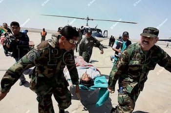 Dos militares de camuflaje corren llevando una camilla con una persona, mientras otros militares y un helicóptero aguardan en una pista de aterrizaje abierta