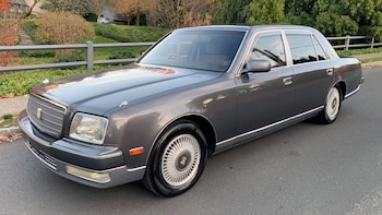 Automóvil sedán Toyota Century de color gris oscuro con llantas plateadas, estacionado en una calle de asfalto junto a una valla y vegetación