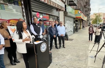 Una mujer habla ante un atril con micrófonos en una acera frente a un comercio, acompañada por varias personas y ante cámaras de televisión