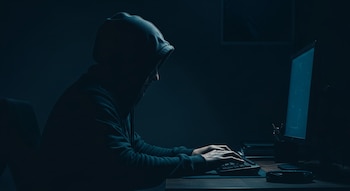 Figura encapuchada sentada frente a un ordenador en una habitación oscura, manos sobre el teclado y pantalla encendida con brillo azul.