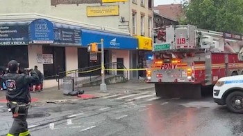 Un camión de bomberos rojo del FDNY y un coche de policía en una calle mojada. Cinta amarilla de precaución rodea un edificio con señales visibles