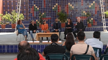 Seis personas sentadas en un panel con micrófono y plantas en macetas, frente a una pared de azulejos de colores y una audiencia de espaldas.