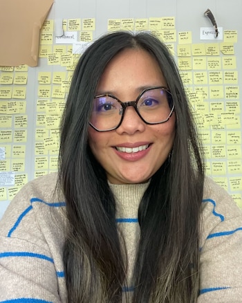 Primer plano de una mujer sonriente con pelo oscuro y gafas. Lleva un suéter beige de cuello alto con rayas azules. Detrás, una pared con notas adhesivas amarillas