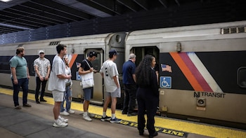 Personas esperan en una plataforma de tren gris y amarilla, frente a un tren plateado de NJ Transit con franjas diagonales naranja, morado y blanco