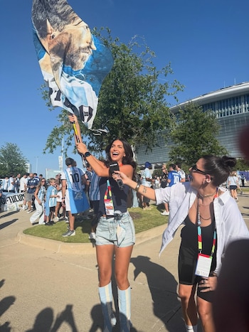 Una mujer sonriente levanta una bandera argentina con el rostro de un hombre barbudo y sujeta un micrófono. Otra mujer sonríe a su lado entre una multitud y un estadio