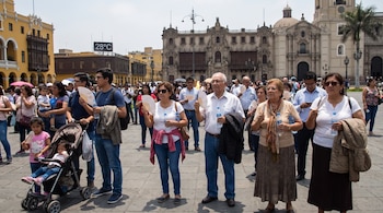Varias personas, incluyendo niños en un coche, en la Plaza de Armas de Lima. Usan abanicos, llevan agua y abrigos. Un cartel indica 28°C.