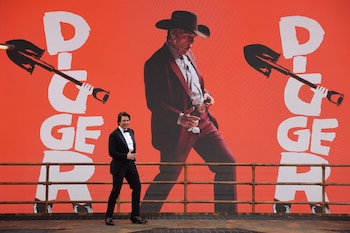 Tom Cruise en esmoquin camina frente a una pantalla naranja con la palabra Digger y una figura masculina con sombrero de vaquero