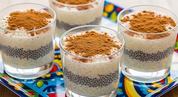 Cuatro vasitos de cristal con capas de arroz con leche, semillas de chía y canela en polvo. Están sobre una bandeja rectangular con patrón de colores vibrantes.