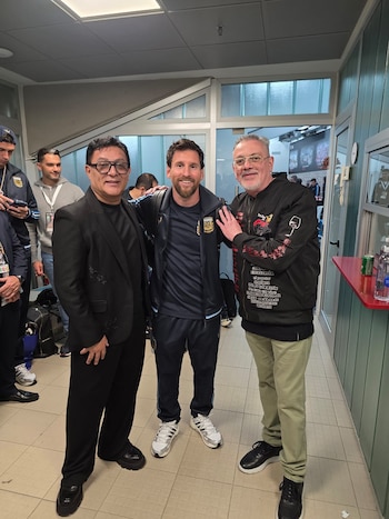 Lionel Messi, vestido con ropa deportiva de la selección argentina, posa sonriente junto a dos hombres en un pasillo