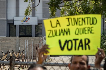 Primer plano desenfocado de una persona sosteniendo una pancarta amarilla con el texto "La juventud zuliana quiere votar". Al fondo, la sede del CNE con vallas