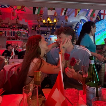 Dos personas sentadas frente a una mesa, una mujer de cabello largo abraza a un hombre que sonríe y sostiene tres rosas rojas. Hay banderas de países en el fondo