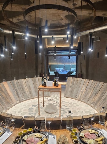 Mesa circular con platos de embutidos, quesos, copas de vino y lámparas colgantes en una sala subterránea con paneles de madera