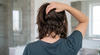 Vista de espaldas de una persona que sostiene con una mano su cabello castaño y deja ver ondas en la zona de la nuca.