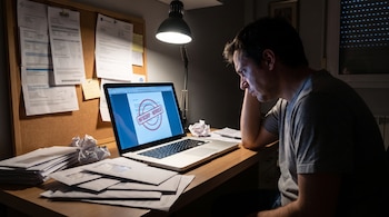 Un hombre de perfil sentado frente a una laptop con un reporte sellado "INFOCORP - MOROSO", rodeado de sobres, papeles arrugados y un tablón con documentos.