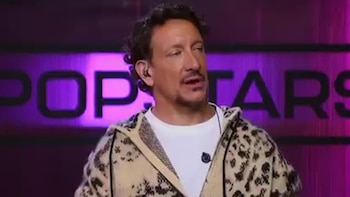 Nicolás Vázquez viste un abrigo estampado y una camiseta blanca frente a un letrero iluminado que dice Popstars