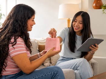 Dos mujeres están sentadas en un sofá; una sostiene una tableta electrónica y entrega una pequeña bolsa de papel rosada a la otra