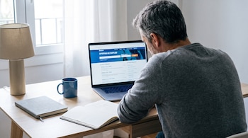Persona de espaldas mirando laptop abierta con ofertas de empleo, en escritorio con taza azul, cuadernos y lámpara, ventana al fondo.