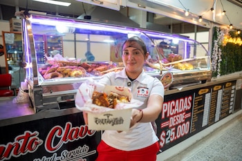 Una mujer detrás de una vitrina iluminada con carne frita extiende una caja con alimentos hacia la cámara