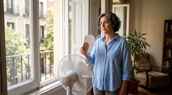 Una mujer con camisa azul sostiene un abanico junto a un ventilador blanco frente a una ventana abierta.