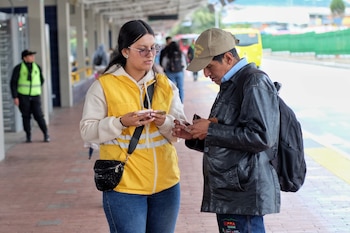 Una mujer con chaleco amarillo de seguridad habla con un hombre con gorra y chaqueta negra que busca en su billetera