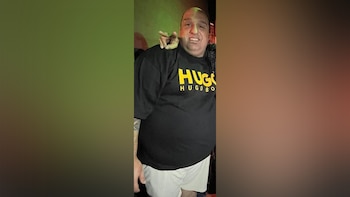 Un hombre calvo con barba, vestido con una camiseta negra con la inscripción "HUGO HUGO BOSS" en amarillo y pantalones cortos blancos, posa sonriendo