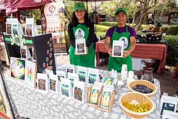 Un hombre y una mujer con delantales verdes sostienen paquetes de café frente a una mesa con productos empaquetados y una cafetera