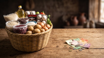 Una canasta de mimbre con granos, aceite, huevos y verduras al lado de varios billetes y monedas sobre una mesa de madera.