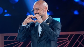 Hombre calvo con anteojos, barba y saco oscuro hace un corazón con las manos en un escenario iluminado con luces azules.