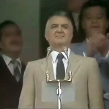 Miguel de la Madrid, de medio cuerpo, en traje claro, camisa y corbata oscura, detrás de un podio con micrófonos. Figuras borrosas de fondo.