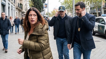 Una mujer con abrigo verde oliva camina por una calle, acompañada por dos hombres. Uno lleva gorra de la Guardia Civil y el otro un auricular. Un vehículo policial es visible.