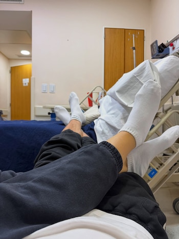 Dos pares de piernas con calcetines blancos y pantalones oscuros sobre camas de hospital con sábanas blancas y azules, en una habitación de clínica