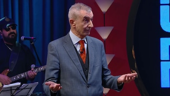Nelson Castro, un hombre mayor con traje gris y corbata naranja, gesticula con las manos abiertas en un escenario. Detrás, un músico barbudo con sombrero toca el bajo