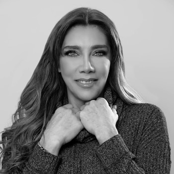 Primer plano de mujer sonriendo, con cabello largo, suéter de cuello alto y manos cruzadas sobre el pecho. Imagen en blanco y negro