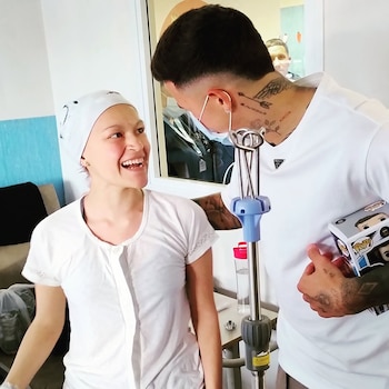 Una mujer sonriente con pañuelo blanco mira a un hombre con tatuajes y mascarilla que sostiene una caja junto a un soporte de suero