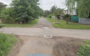 Esquina de una calle pavimentada y otra de tierra con casas, vegetación, tendido eléctrico y un cartel vial