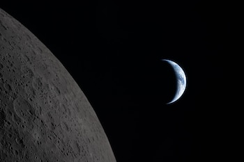 La Tierra en forma de media luna, azul y blanca, flota en la distancia sobre un fondo negro, junto a la superficie gris y craterizada de la Luna en primer plano