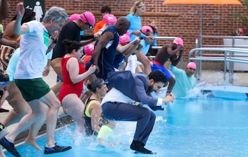 Varias personas saltan a una piscina. Un hombre con traje formal y calcetines entra al agua. Muchos usan gorros de natación rosas. Hay una pared de ladrillos