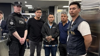 Cinco hombres de pie en un espacio interior, entre ellos oficiales con chalecos de seguridad policial y aeroportuaria